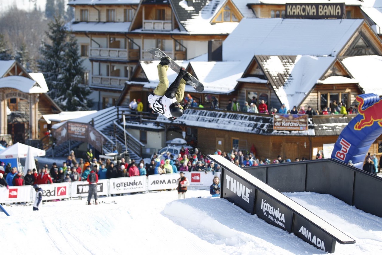 Snowboardzista w powietrzu nad railami, w tle widownia i góralska karczma